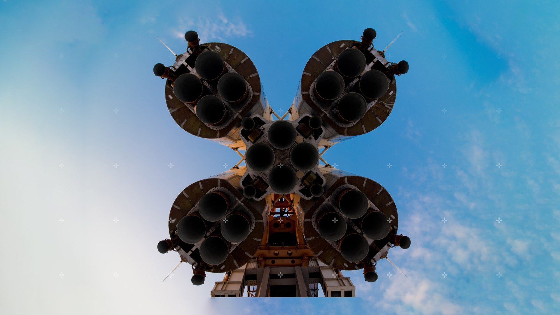 The underside of thrusters for a large rocket, framed against a bright blue sky with a smattering of clouds. A neat row of small, digital-looking crosses are superimposed on top of the image.